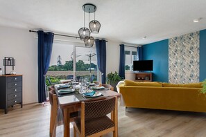 Un comedor con una mesa dispuesta para cuatro personas, un sofá, un televisor y vistas al exterior.