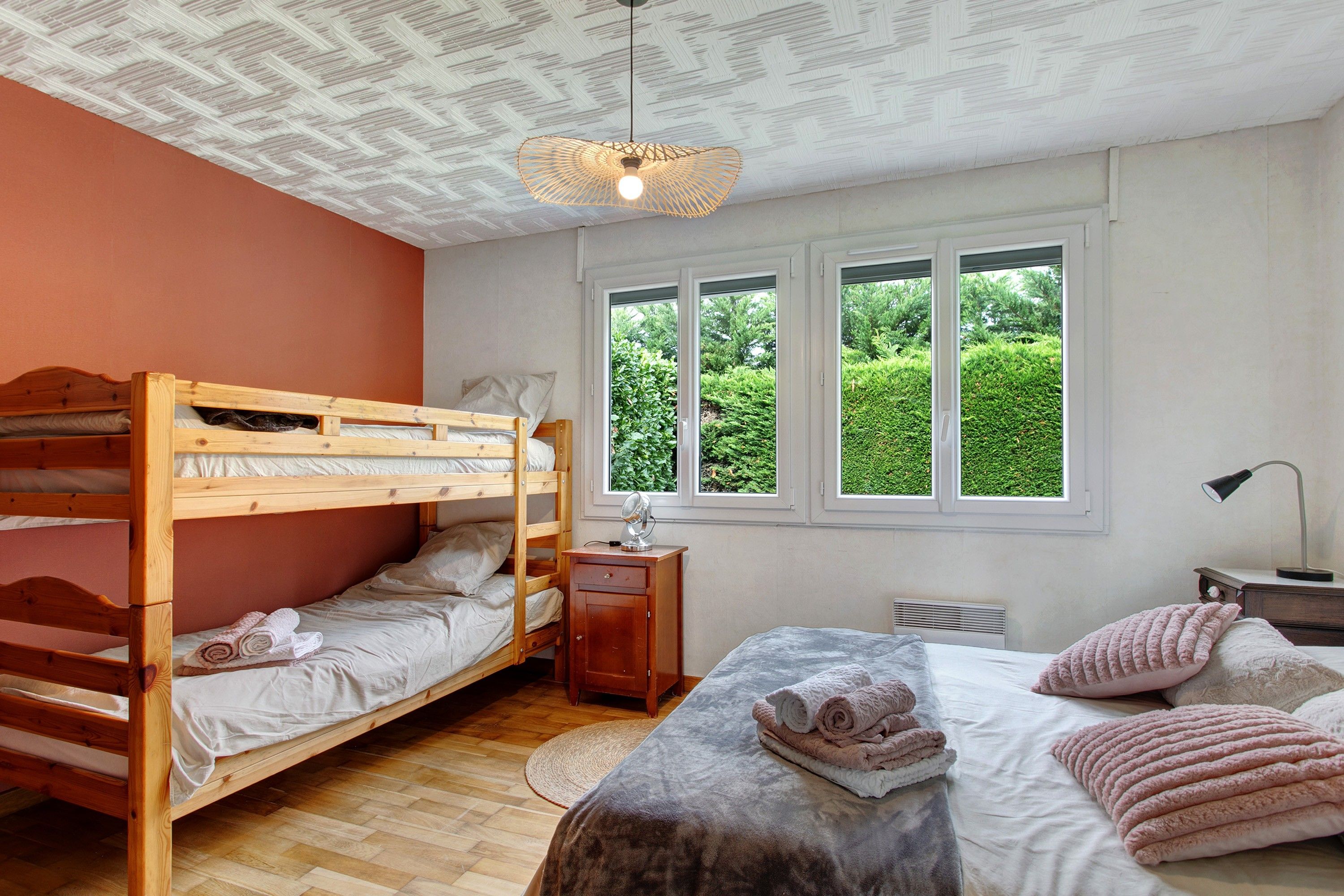 Un dormitorio con literas, una cama individual, una mesita de noche con lámpara, un ventanal con vistas a zonas verdes y un techo con un diseño original.