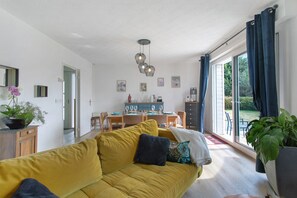 Un salón luminoso con un sofá amarillo, comedor y ventanas amplias con cortinas.