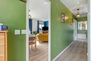A hallway with green walls, wooden flooring, a dining area with a yellow sofa, and a TV in the background. - Holiday home 'Petite Maison de Kervenou' with private terrace, garden and Wi-Fi (Goudelin)
