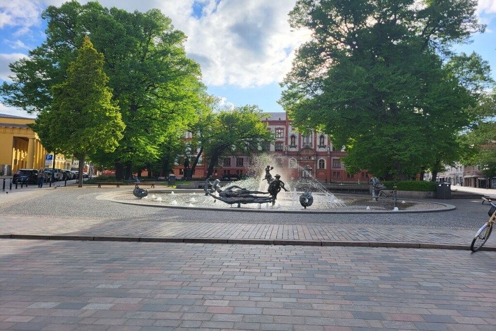 Ein öffentlicher Platz mit einem zentralen Brunnen, umgeben von Bäumen und Gebäuden.