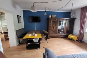 Ein Wohnzimmer mit Esstisch, einer Couch, einem an der Wand befestigten Fernseher, einem Holzkasten und einer Treppe.