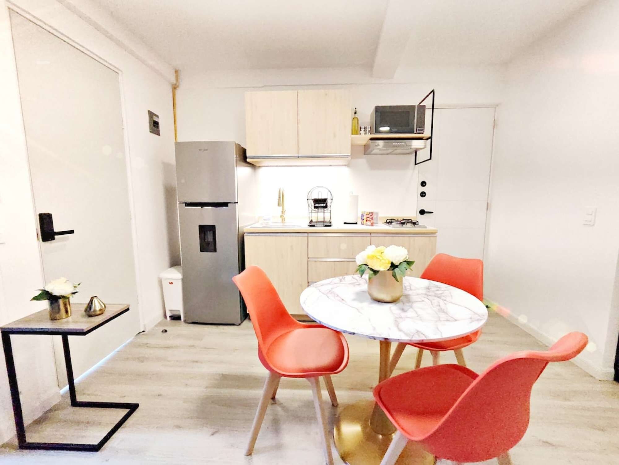 Une cuisine compacte équipée d’un réfrigérateur, d’un four à micro-ondes et d’une cuisinière à gaz. On y trouve une table à manger avec deux chaises et un vase rempli de fleurs.