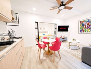 Une cuisine moderne dotée d’une cuisinière intégrée, un coin repas avec une table ronde et des chaises rouges, un ventilateur de plafond, un téléviseur fixé au mur et un tableau abstrait aux couleurs vives.