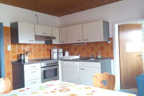 Eine kleine Küche mit Herd, Backofen und Kaffeemaschine, ein Esstisch mit Blumenmuster bedecktem Tuch und ein Holzstuhl mit Herzförmigem Ausschnitt.