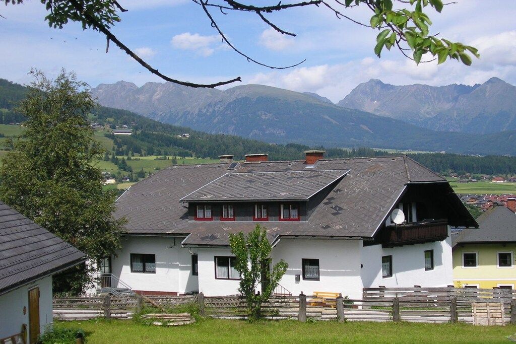Un bâtiment blanc avec un toit en pente, un petit arbre devant et une chaîne de montagnes en arrière-plan.