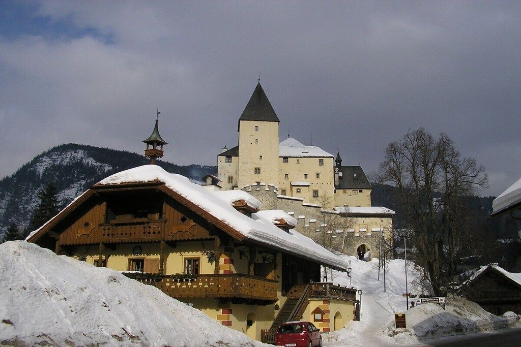 A traditional chalet with a balcony, a tower, and a castle-like building in the background.