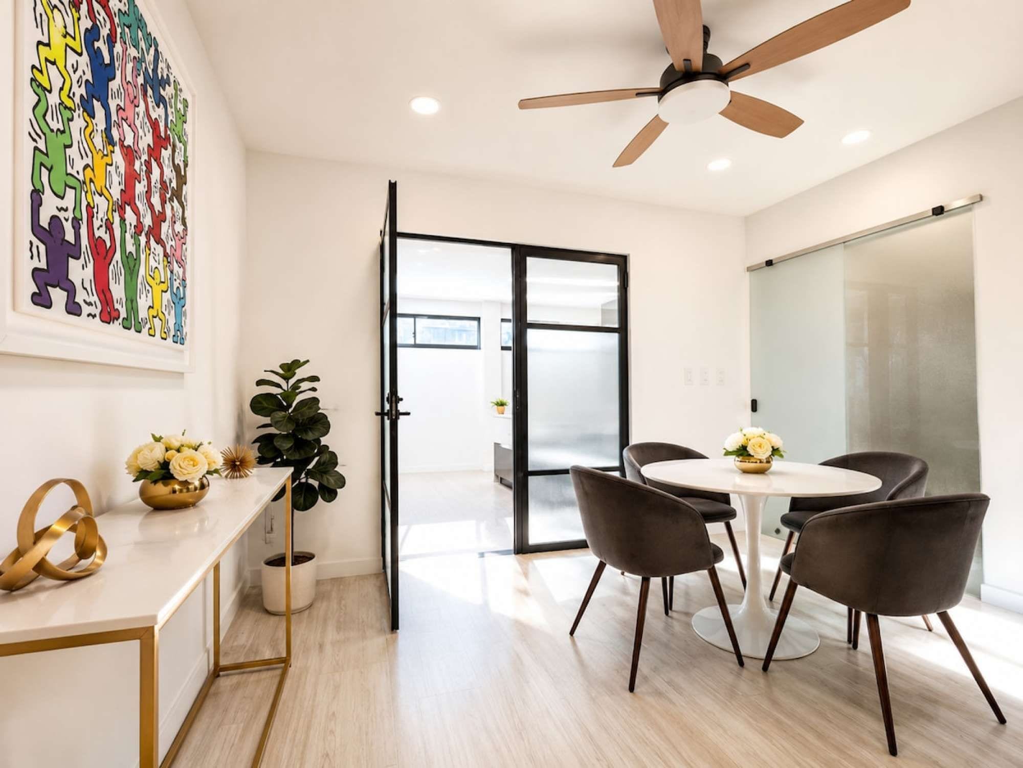 A modern office space with a round table, black chairs, a ceiling fan, and a glass door leading to another room.
