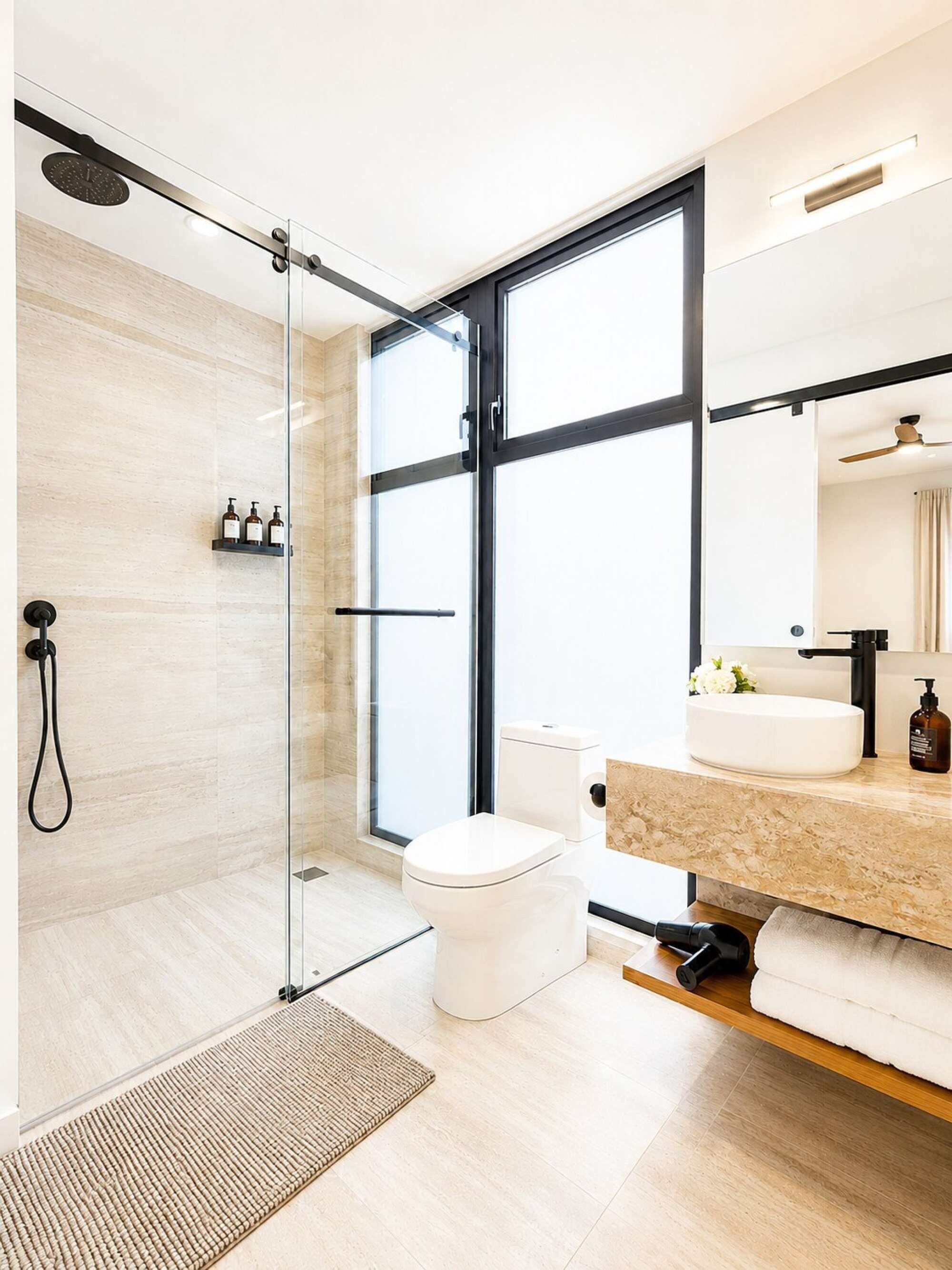 A modern bathroom with a glass shower enclosure, a toilet, a sink with a vanity, and a large window providing natural light.