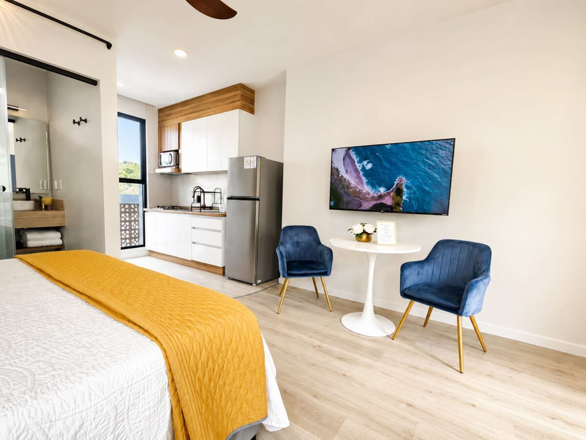 A modern hotel room with a bed, a dining area with two chairs, a kitchen, and a large TV on the wall.