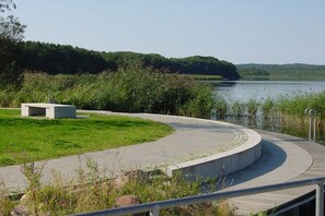 Ein gewundener Betonweg führt zu einem Gewässer, umgeben von Grünpflanzen und einer kleinen Betonkonstruktion.