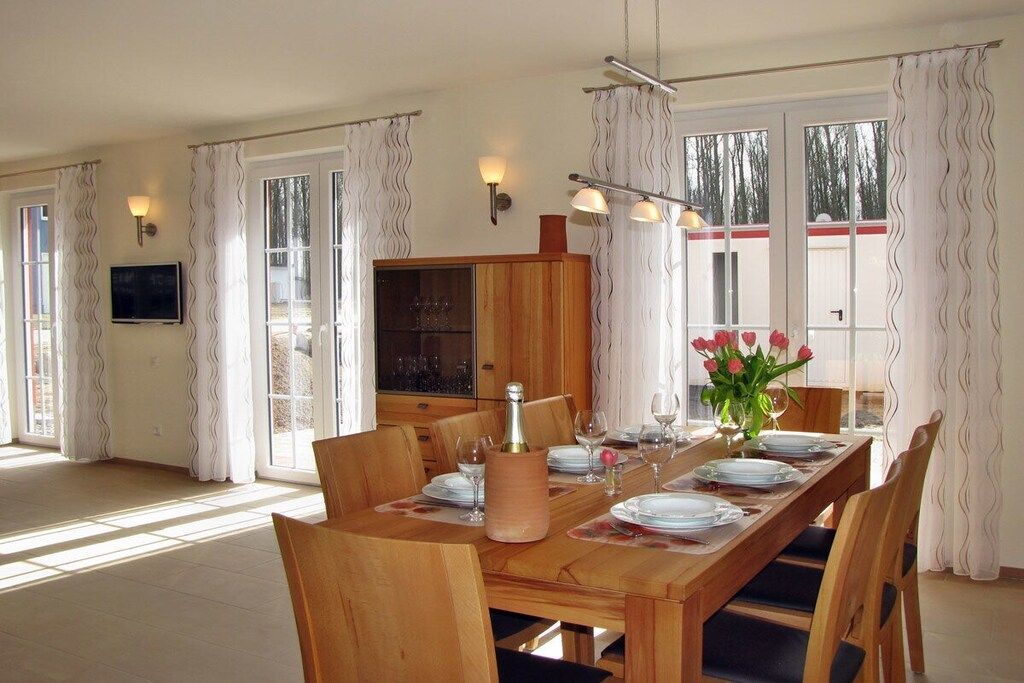 Un coin repas avec une table en bois dressée pour quatre personnes, bordé de rideaux et agrémenté d’un téléviseur fixé au mur.