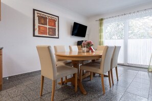 A dining area with a round table, four chairs, a flat-screen TV mounted on the wall, and a window with curtains. - House Hill Apartment 3 (Ostseebad Binz)