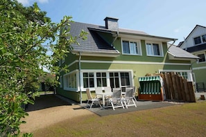 Ein zweistöckiges Haus mit überdachter Terrasse und einer kleinen Sitzgruppe im Freien.