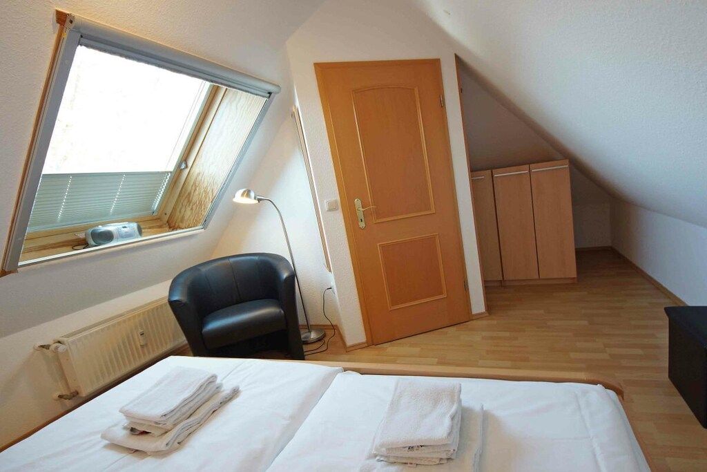 A small, single-room apartment with a sloped ceiling, a window, a door, a radiator, a chair, and a bed with neatly folded white towels.