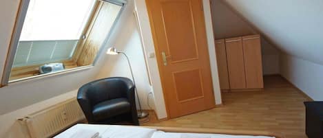 A small, single-room apartment with a sloped ceiling, a window, a door, a radiator, a chair, and a bed with neatly folded white towels.