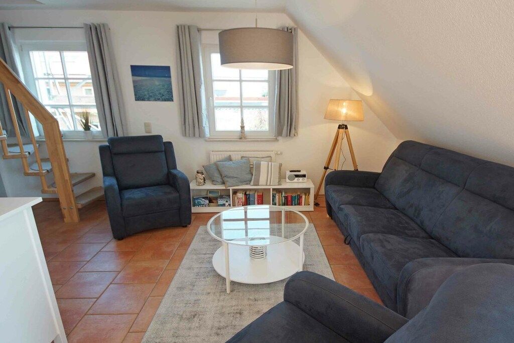 Ein Wohnzimmer mit einer Couch, einem Sessel und einem Couchtisch. Ein Bücherregal mit Büchern und einer Lampe sind ebenfalls zu sehen. Ein Fenster mit Vorhängen ist erkennbar, und im Hintergrund befindet sich eine Treppe.