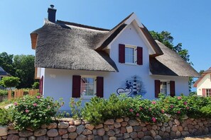 Ein traditionelles Haus mit einem Strohdach, einer steinernen Mauer davor und der Hausnummer 98 an der Seite.