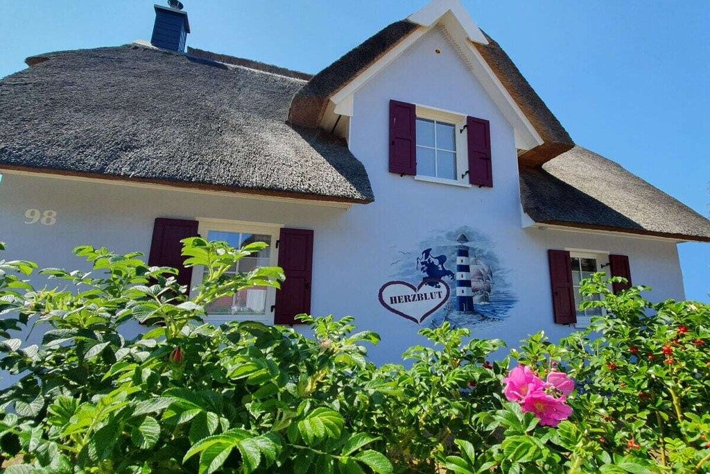 A white building with a thatched roof and red shutters, marked with the number 98 and a heart-shaped logo with the text 'HERZBLUT'.
