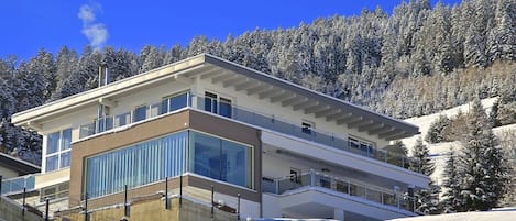 A modern multi-story building with large windows and balconies, surrounded by a snowy landscape and trees.