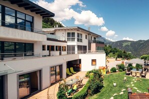 Ein modernes, zweistöckiges Haus mit einem Balkon, einer Terrasse mit Gartenmöbeln und Blick auf die Berge.