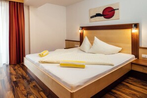Ein ordentlich gemachtes Bett mit Kissen und einem zusammengerollten Handtuch, ein Holz-Kopfteil und ein an der Wand hängendes Kunstwerk.