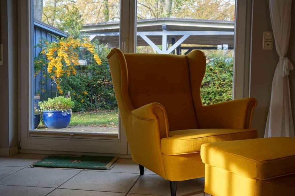 Una habitació amb una butaca groga i un reposapius, una finestra amb vista a un jardí, i una porta blava.