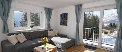 A modern living room with a grey sofa, a wooden coffee table, and a view of mountains through large windows.