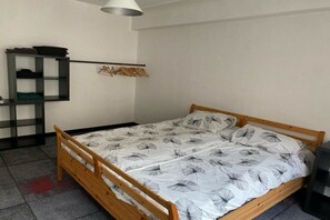 Ein Einzelbett mit Holzrahmen und weißer Bettdecke mit Blätermotiv, ein kleines Regal mit gefalteter Kleidung und ein an der Wand befestigter Wäscheständer mit Kleidern.