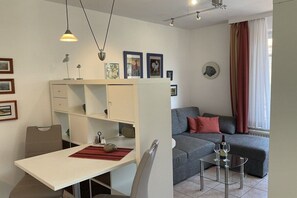 Ein modernes Wohnzimmer mit einer Couch, einem Esstisch und einem Glastisch. An der Wand befinden sich Bilder in Rahmen und ein Fenster mit Vorhängen.