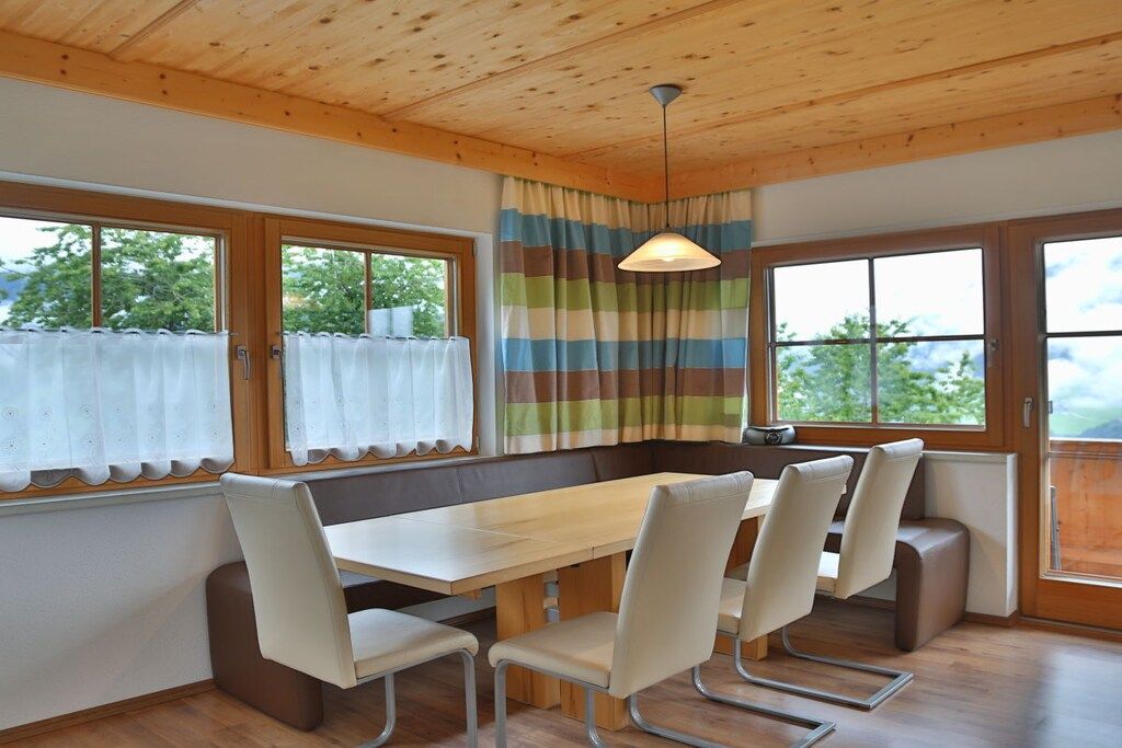 A dining area with a wooden table and chairs, large windows with curtains, and a view of greenery outside.