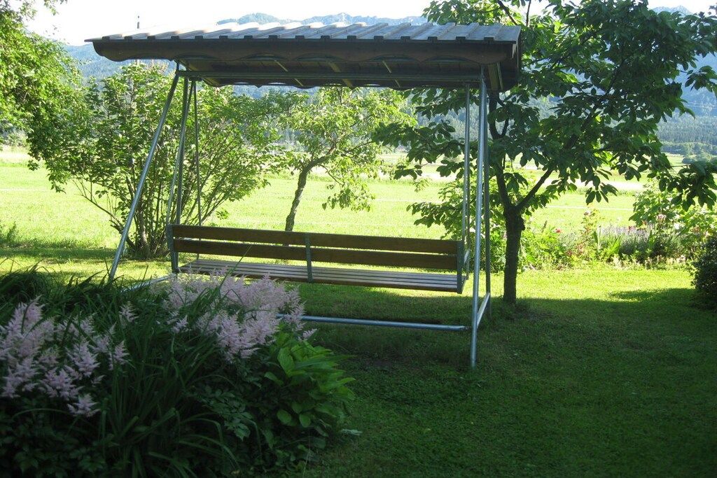 A wooden bench under a metal pergola in a grassy area with trees and a mountain view.