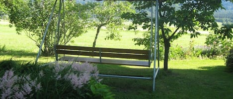 A wooden bench under a metal pergola in a grassy area with trees and a mountain view.