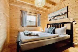 Ein Zimmer in einer Holzhütte mit einem Bett, einem Vorhangfenster, einem Gemälde an der Wand und einer Lampe.