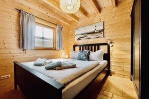 A wooden cabin bedroom with a bed, a window with curtains, a painting on the wall, and a nightstand with a lamp. - Log cabin Seagull's nest (Dierhagen)