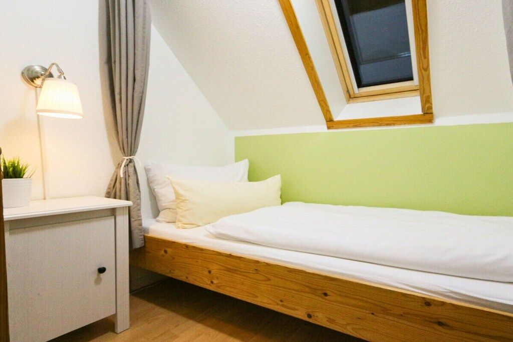 Ein Einzelbett mit Holzrahmen und einem grünen Kopfteil, ein Nachttisch mit Lampe und ein Fenster mit Holzrahmen.
