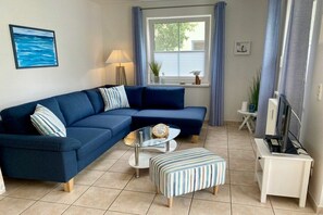 Ein Wohnzimmer mit einem blauen Sofa, einem gestreiften Hocker, einem Glastisch und einem Fernseher auf einem Schrank.