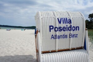 A beach chair labeled 'Villa Poseidon Atlantis Binz' on a sandy beach with a view of the sea and distant hills. - Villa Poseidon 14 - Atlantis Binz (Ostseebad Binz)