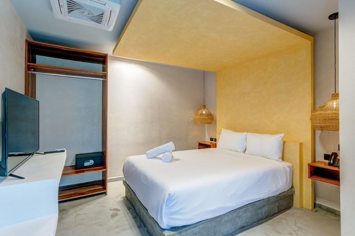 Quarto de hotel com cama, televisão, guarda-roupa e mesa de cabeceira com abajur.