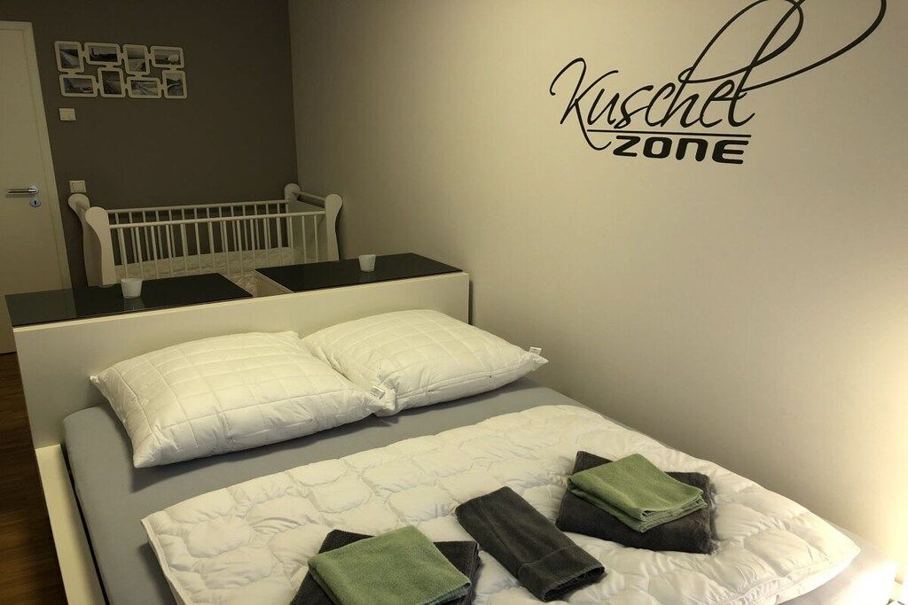 Une chambre avec un lit blanc, deux oreillers, un berceau et un panneau mural sur lequel on peut lire : « Zone câlins ».