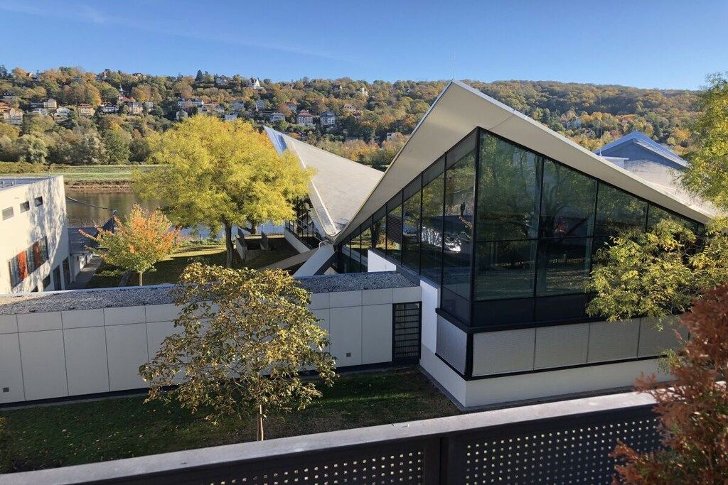 Un bâtiment moderne doté d’un toit en pente et de grandes fenêtres en verre, entouré d’arbres et avec une colline en arrière-plan.