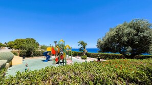 A playground with slides and climbing structures, a bench, and a view of the sea.