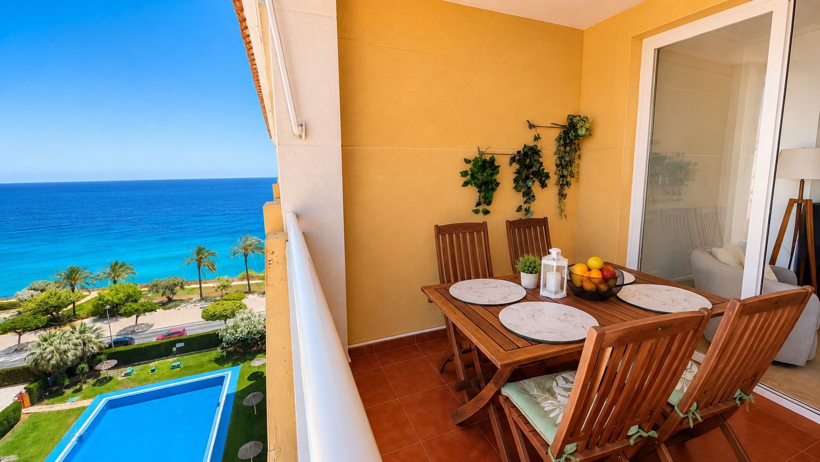 A balcony with a wooden table set for two, a bowl of fruit, and a view of the ocean.