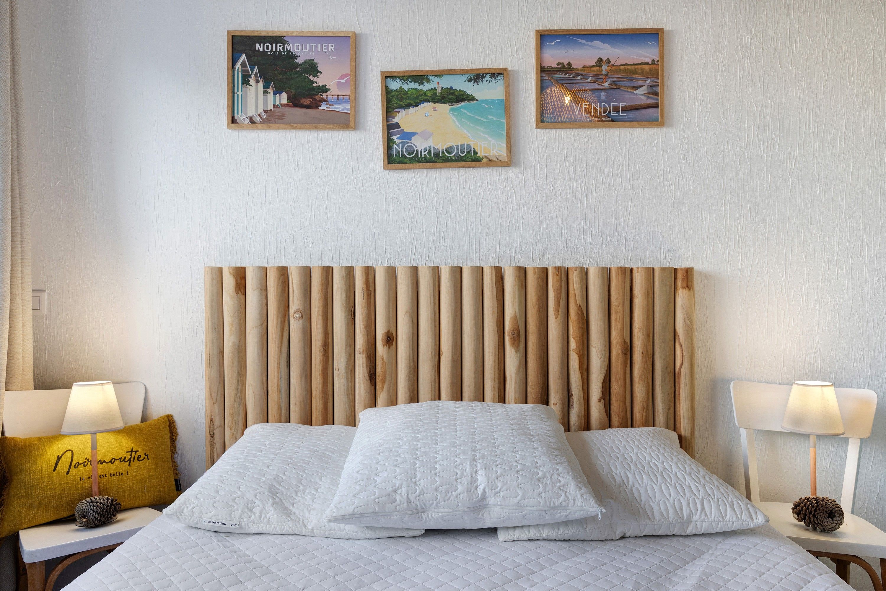 Une chambre d’hôtel avec une tête de lit en bois, un lit recouvert de draps blancs, deux tables de chevet avec des lampes et trois tableaux encadrés accrochés au mur.