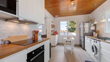Una cocina moderna con suelo de madera, muebles blancos, horno empotrado, lavadora y comedor con mesa y sillas.