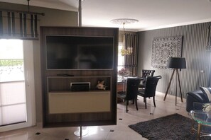 A modern living room with a flat-screen TV, a dining area with a table and chairs, and a black floor lamp. - Holiday home with 1 bedroom (Fuhlendorf)