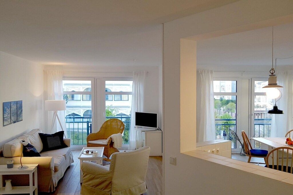 Ein modernes Wohnzimmer mit einer Couch, einem Esstisch und einem Fernseher. Große Fenster mit Vorhängen, ein Balkon und eine Aussicht auf Gebäude.
