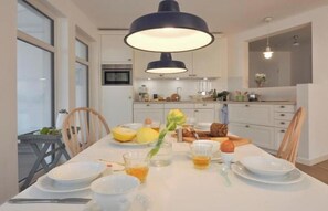 A dining area with a white table set for four, a modern pendant light, and a kitchen in the background. - Villa Louisa 47 - Montauk (Ostseebad Sellin)