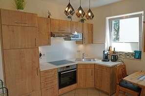 Une cuisine compacte avec des meubles en bois, un four encastré, un four à micro-ondes et un évier. Des luminaires suspendus sont placés au-dessus du plan de travail, et une fenêtre apporte de la lumière naturelle.