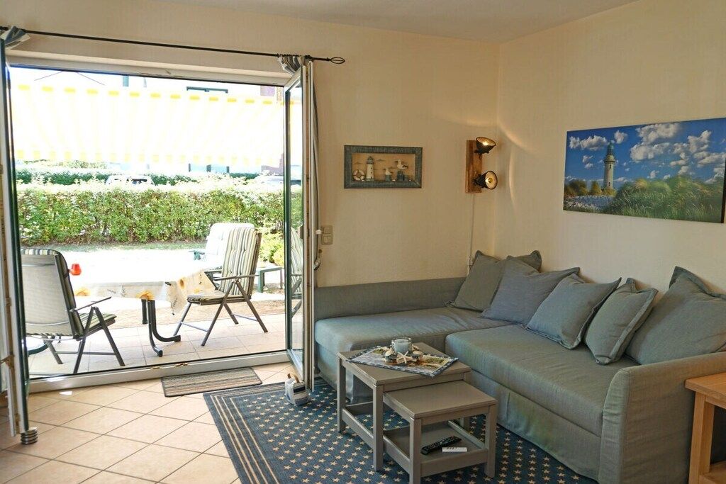 Ein Wohnzimmer mit einer Couch, einem Couchtisch und einem Sessel. Eine Tür führt zu einem Außenbereich mit Blick auf Grünflächen und ein Gebäude.